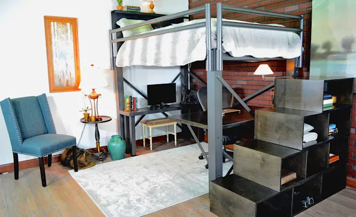 Stylish room with a loft bed above a study area featuring a desk, chair, and computer. A staircase with storage compartments leads to the bed. There's an upholstered chair, lamp, framed artwork, and decor elements on a wooden floor with a brick accent wall.