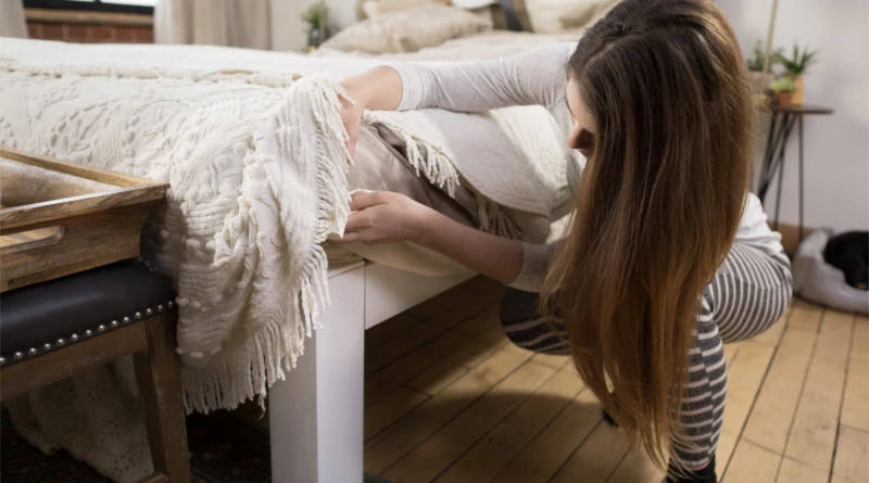 A woman finishes making her white Queen Standard Bed. She is kneeling at the foot of the bed and lifting the comforter, unveiling all of the extra space under the bed frame for storage.