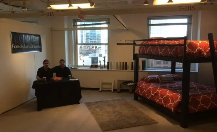 A showroom features a tall bunk bed with red patterned bedding. Two people sit at a table with a black tablecloth. The room has large windows and overhead lights, and a rug is placed on the floor. A sign reads "Francis Lofts & Bunks.
