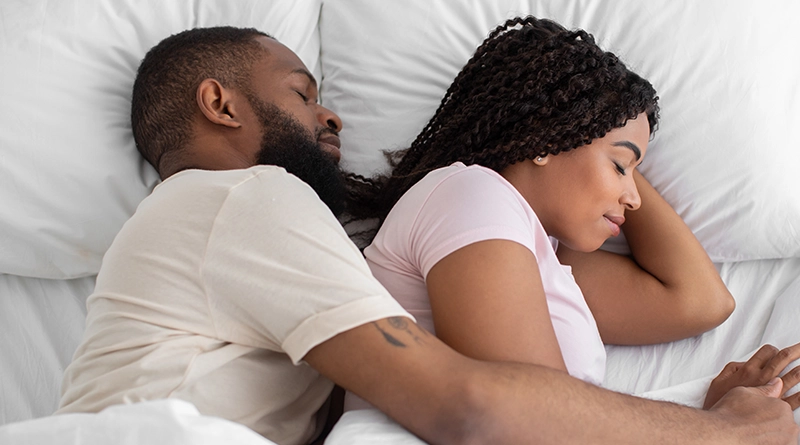 Young Black Couple Lying in Bed together with man wrapping his left arm around the woman while they sleep - 800x445