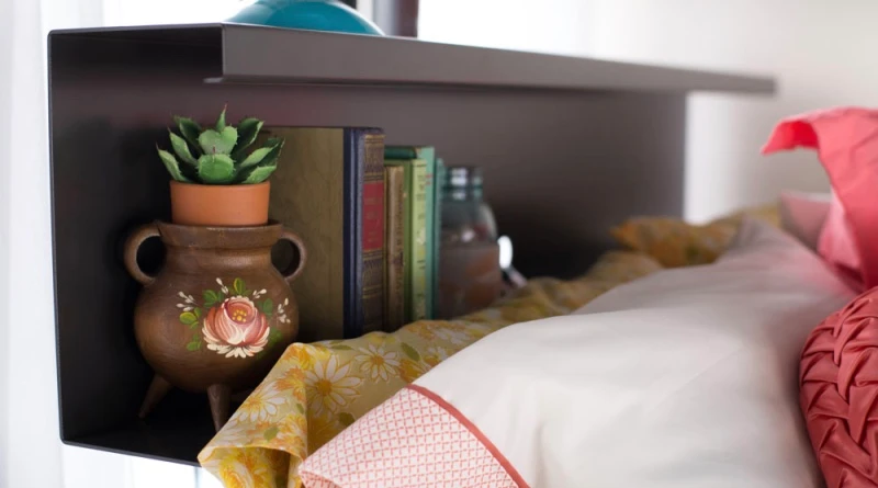 A close-up of a decorative headboard shelf featuring a potted plant, several books, and a candle. The bed below is covered with patterned pillows and a floral duvet, creating a cozy and inviting atmosphere.