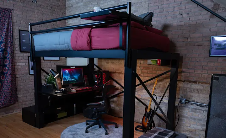 Black Adult Loft Bed in an urban studio apartment with red and gray bedding with an attached black desk an full PC gaming setup on the desk, seen from the lower left-hand corner of the bed frame looking under the sleeping platform.