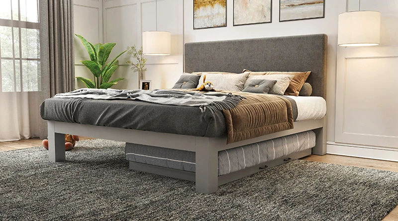 A light gray colored King size metal Platform Bed with a dark gray headboard and Twin size metal trundle tucked underneath the left-hand side of the bed. Seen here from the lower left-hand corner of the bed on the side of the trundle.