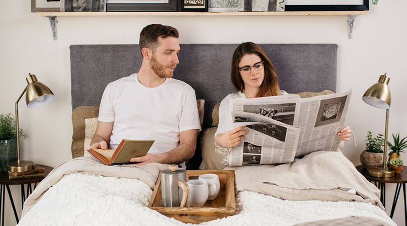 A young couple sits propped against the headboard of their white platform Standard Bed reading a book and newspaper, respectively, and talking to one another.