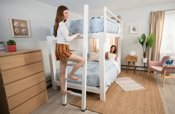 Two young women in a beach house bedroom with a white Queen Over Queen Adult Bunk Bed. One is lying on the bottom bunk under the covers while the other climbs up the ladder at the foot of the bed.