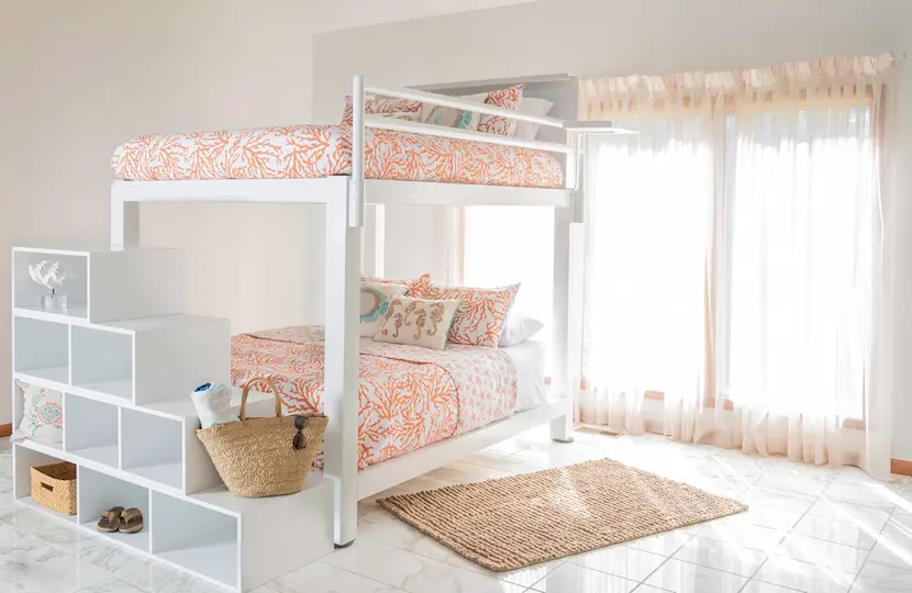 A white bunk bed for adults with a matching wooden staircase and orange and white patterned bedding in a sparsely decorated guest room.