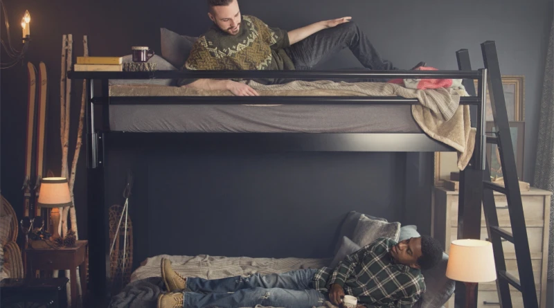 A Black Queen Over Queen Adult Bunk Bed with a Man on Each Bunk. The men are looking at each other from their bunks and talking. The top bunk has a tray accessory with books and a coffee mug. The bunk bed is located in a ski resort bedroom.