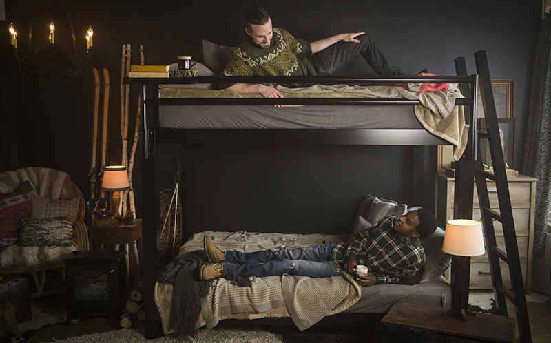 Two adult men lying on each bunk of a black Queen Adult Bunk Bed in a ski resort bedroom looking at each other and talking from their bunks - 800x498