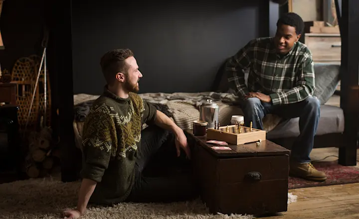 Two young adult men sit talking. One is sitting on the bottom bunk of a black Queen Over Queen Adult Bunk Bed. The other is sitting on the floor next to him. There is a coffee table with a chess board between them.