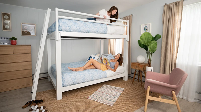 Two young adult women lying on both bunks of a white Queen Over Queen Adult Bunk Bed and talking to one another in a beach house vacation rental