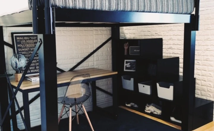 A loft bed with a striped mattress sits above a workspace featuring a wooden desk, clear chair, and a shelving unit with storage bins. The space is set against a white brick wall and light wood flooring, creating a modern, organized room.
