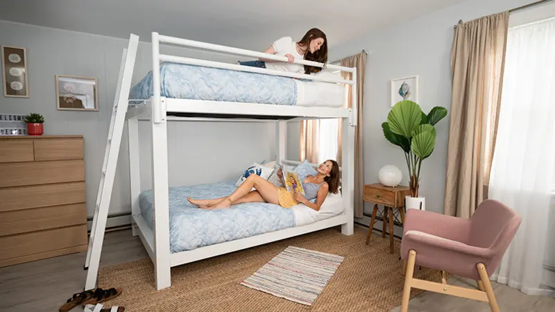 Two young women on each bunk of a white Adult Bunk Bed talking to one another by looking down or up, respectively. The bunk bed is in a bright and airy beach house vacation rental. 