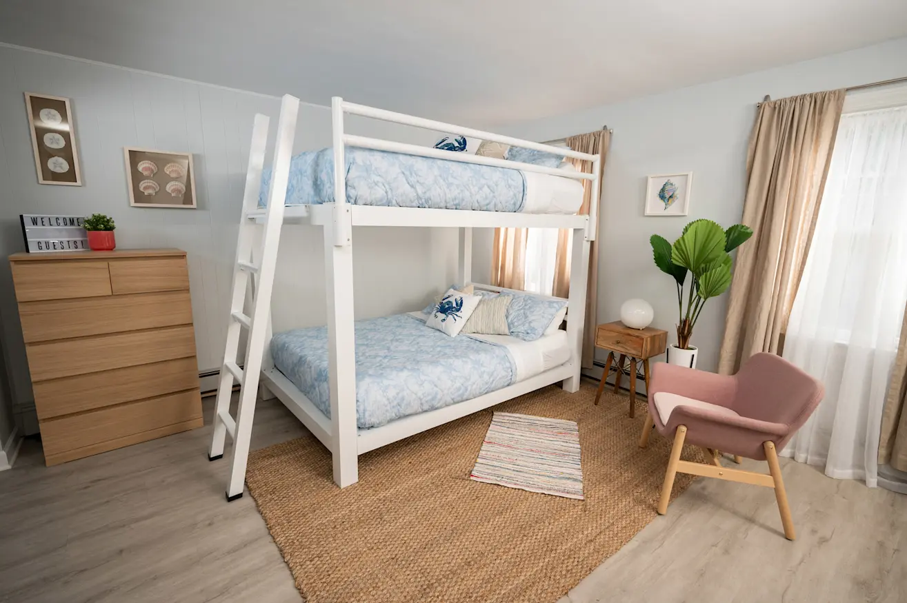 A cozy bedroom with a white bunk bed, blue bedding, a wooden dresser, and a pink chair. A potted plant sits by the window, and a small rug is on the floor. Neutral walls are decorated with framed art.