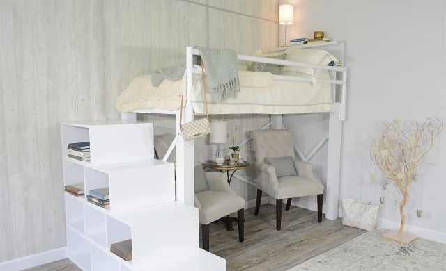 A wide corner angle view of a full size white Adult Loft Bed with a staircase, bookshelf, and two grey chairs beneath the bed. 