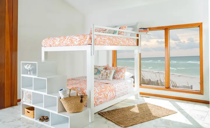 A bright bedroom with a white loft bunk bed featuring orange-patterned bedding. The bottom bunk is a queen-sized bed. There's a shelf-staircase with a basket and decor items. A large window offers a view of the ocean and beach.