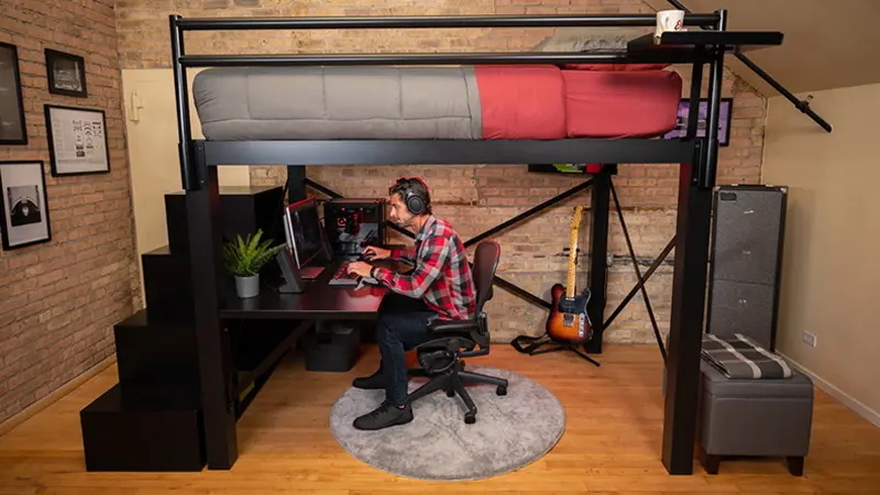 An adult male gamer playing on his computer underneath his black Queen Adult Loft Bed.
