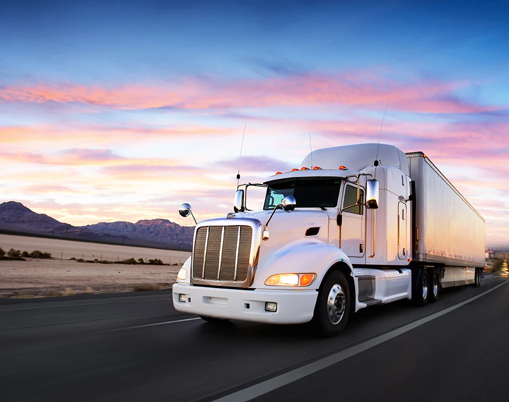 A white semi truck with an attached can hauling items down an empty highway road at dusk.