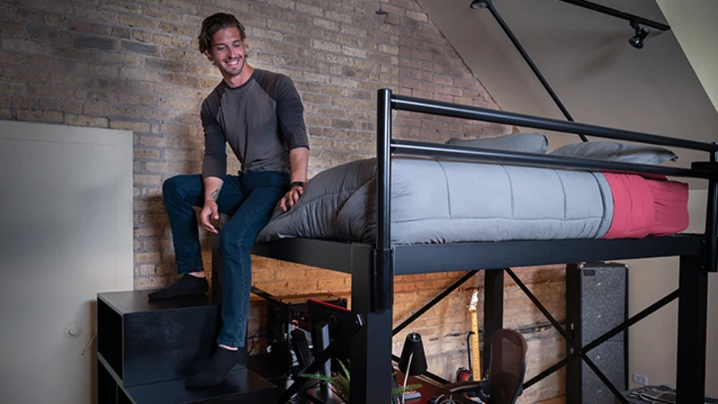 Adult man sitting at the foot of a black queen size Adult Loft Bed with his feet hanging off the end of the bed and resting on the matching wooden staircase.
