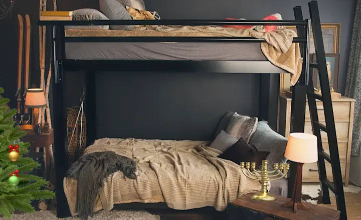 A cozy room with a loft bed, ladder, and assorted blankets. Below, the lower bed is surrounded by pillows. A menorah and table lamp sit on a nearby chest. Skis are propped up against the wall. A small decorated Christmas tree is visible.