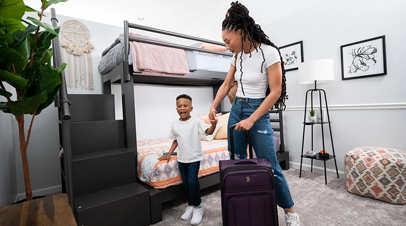 A young mother with her young son standing in a guest room with a charcoal Twin XL Over Queen Bunk Bed for adults with a staircase holding a rolling suitcase.