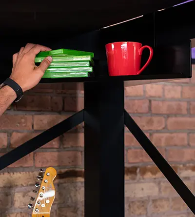 A man reaching for a video game he has sitting on his Corner Shelf accessory under his black Queen Size Adult Loft Bed.