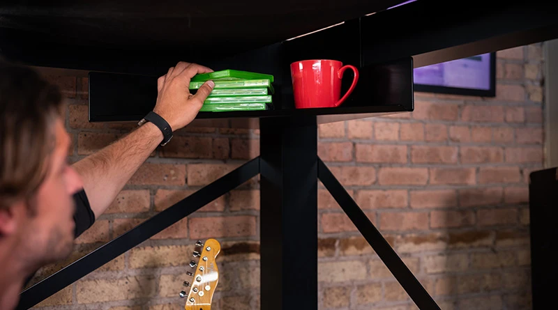 A man reaching for a video game he has sitting on his Corner Shelf accessory under his black Queen Size Adult Loft Bed.