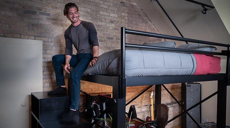 A large adult man sitting at the foot of his black Queen Adult Loft Bed with his feet resting on the matching staircase in a studio apartment with exposed brick walls.