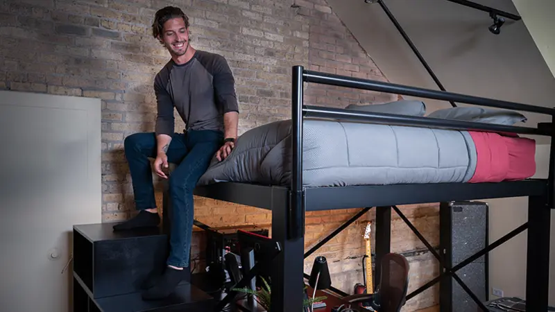 A large adult man sitting at the foot of his black Queen Adult Loft Bed with his feet resting on the matching staircase in a studio apartment with exposed brick walls.