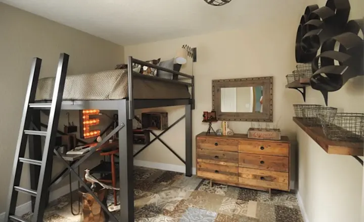 Bedroom with a loft bed featuring a metal frame. A desk and chair are underneath. The room is decorated with rustic furniture, including a wooden dresser and shelves. The floor has a patterned rug, and a metal light fixture hangs from the ceiling.