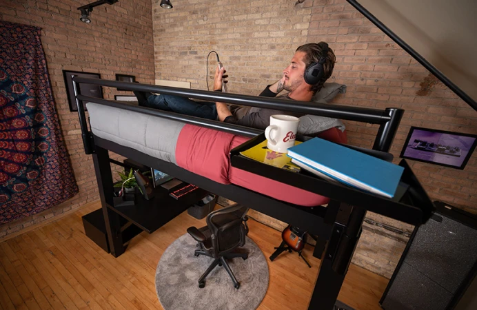 Adult man lying on a black Adult Loft Bed listening to music in headphones and seen from the side of the bed at a slight angle looking down over his left shoulder.