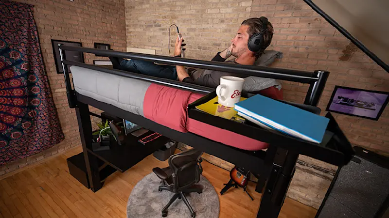 Adult man lying on a black Adult Loft Bed listening to music in headphones and seen from the side of the bed at a slight angle looking down over his left shoulder.