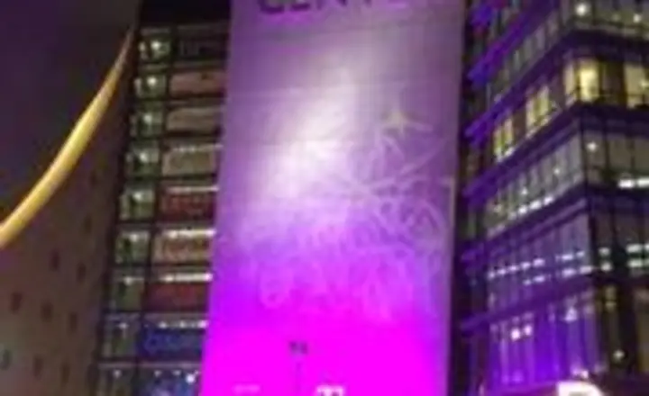 A night scene at the World Market Center. The tall building is illuminated in purple, surrounded by other lit-up structures. People gather outside, some carrying glowing items, adding to the vibrant atmosphere.