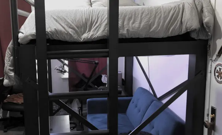 Lofted bed with gray bedding above a blue sofa in a compact room. Red curtains cover the windows, and a small desk with a microphone setup is visible. Gray and black accents complete the modern layout.