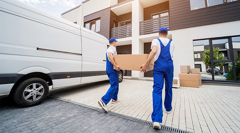Two strapping delivery men in white t-shirts, blue overalls, and blue hats both holding each end of a long cardboard box and walking it into a condo building. Seen from behind next to their white van.