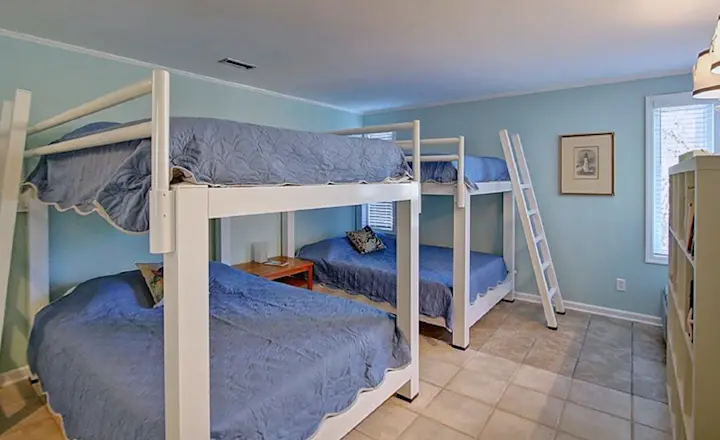 A bedroom with two sets of white wooden bunk beds, each with blue bedding. A small wooden table is between the beds. The room has a pale blue wall, tiled flooring, a window with blinds, and a lamp on a shelf in the corner.