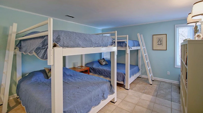 A bedroom with two sets of white wooden bunk beds, each with blue bedding. A small wooden table is between the beds. The room has a pale blue wall, tiled flooring, a window with blinds, and a lamp on a shelf in the corner.