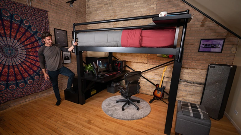 A man stands next to a modern loft bed with gray and red bedding. Below is a desktop setup with a chair and a guitar leaning against the desk. The room features exposed brick walls, a tapestry, and framed art.