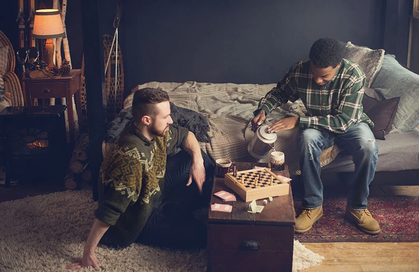 Two men in a ski lodge vacation rental bedroom with a black Queen Over Queen Adult Bunk Bed. One sits on the floor next to a table with a chess board while the other sits on the bottom bunk and pours coffee into a mug.