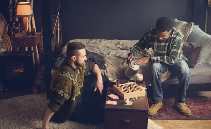 Two people sitting in a cozy room with a bed, playing a board game on a small table. One is pouring tea from a kettle, and the other is sitting on the floor. The room is warmly lit with a rustic and comfortable decor.