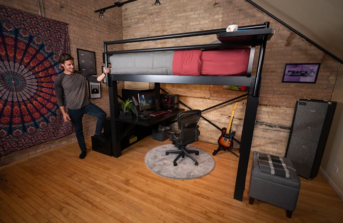 Black Queen Loft Bed for Adults in an urban studio apartment with brick exposed walls with a young adult man standing at the foot of the bed with his foot on the bottom step of the matching wooden staircase.