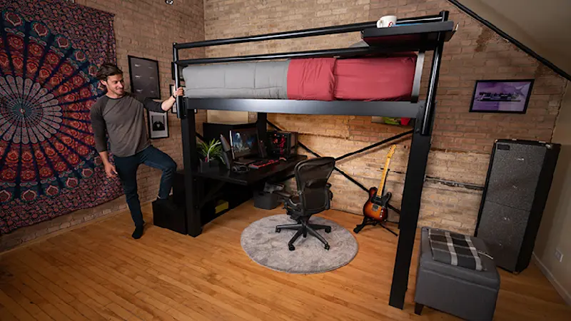 Black Queen Loft Bed for Adults in an urban studio apartment with brick exposed walls with a young adult man standing at the foot of the bed with his foot on the bottom step of the matching wooden staircase.