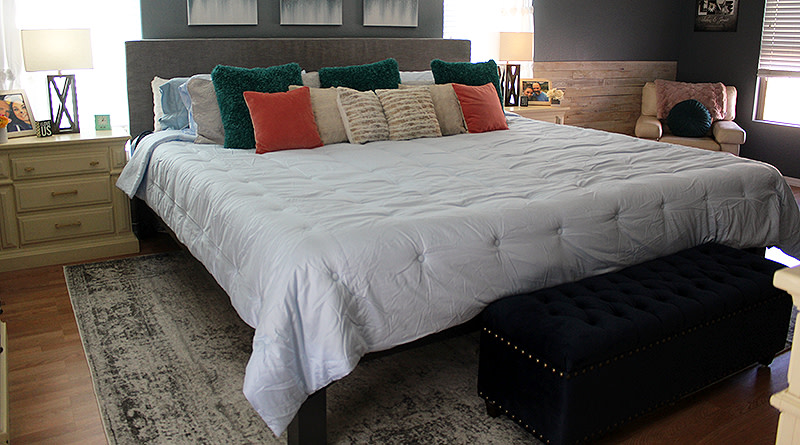 A black Alaskan King size bed frame in a large bedroom with blue walls. The bed has a grey headboard and is covered with a large white comforter and turquoise and orange throw pillows.