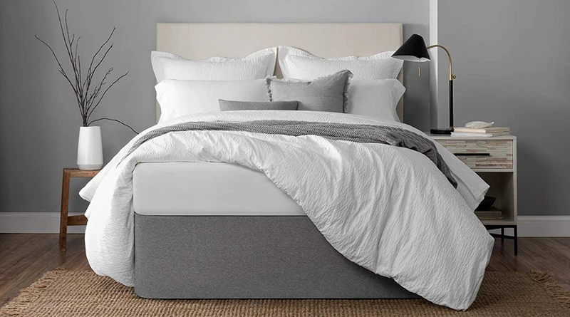 A light gray fabric bed wrap on a platform bed frame with a cream-colored headboard. Bedding is mostly white with a gray throw blanket and pillow.