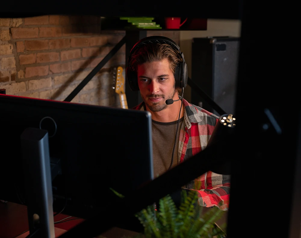 Adult man sitting underneath a queen size black Adult Loft Bed at his attached desk looking at a computer monitor, seen from the side of the bed through frame behind the monitor.