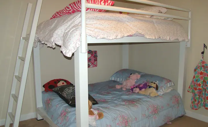 A white bunk bed with a floral-patterned comforter on the upper and lower beds. The top bunk is accessed by a ladder. Plush toys and pillows are on the lower bed. A dress hangs on the wall to the right.