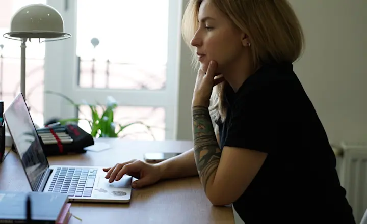 A woman with blond hair sits at a wooden desk, focused on a laptop. She rests her chin on her hand, with a tattoo visible on her forearm. The room is bright with a window in the background and a desk lamp nearby.
