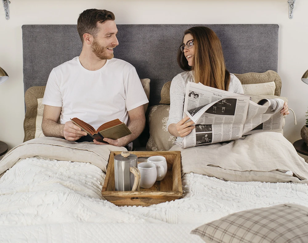 A young couple sitting up in their white queen size metal Platform Bed with a blueish gray headboard. She is reading a newspaper and he is reading from a book while they talk and look at each other. Seen from head-on.
