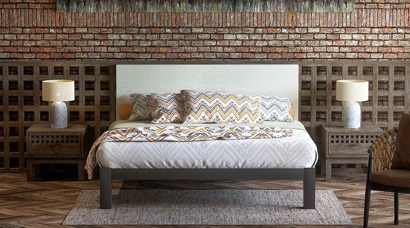 A charcoal Wyoming King Bed with a cream headboard seen from the front in a rustic hotel room with a brick wall background.
