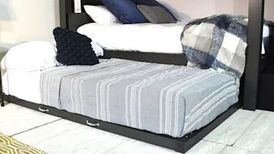 A bedroom setup featuring a bunk-style bed with a lower trundle. The top bed is adorned with white and blue pillows, while the trundle has a gray striped blanket. A plaid blanket is draped over the side. Light wood flooring is visible.