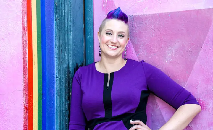 A photo of Dr. Liz Powell, sexy psychologist, standing in front of a purple wall. 